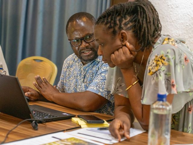 A man sits at a table in front of a laptop. A woman, crouching next to him with her elbows on the table,  is speaking to him.