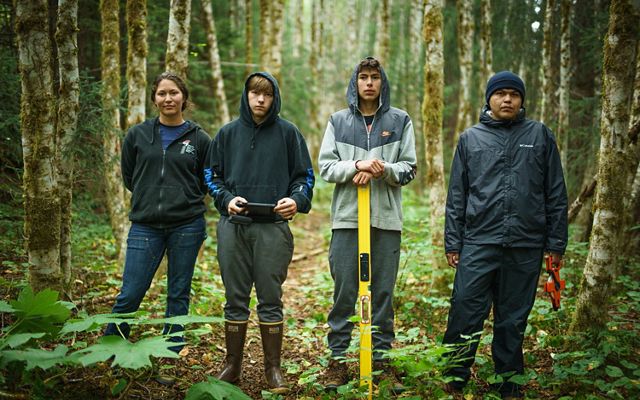 4 young adults wearing boots and jackets stand in a forest while holding various tools.