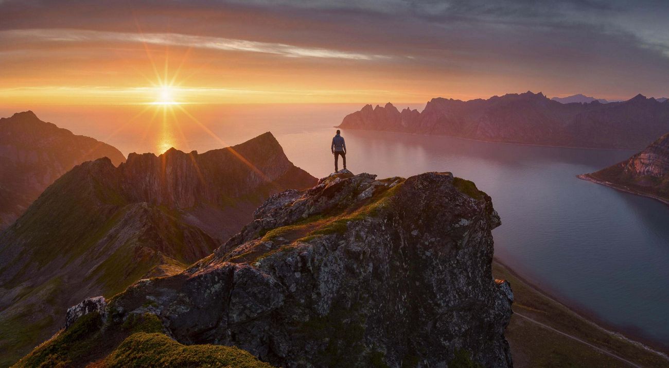 A person stands atop a cliff overlooking a fjord with the midnight sun shining over the sea in the distance.