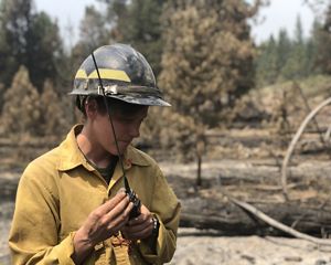 TNC Oregon Fire Program Director Katie Sauerbrey stands in a forest and listens to a walkie talkie; she is wearing fire-protective clothing and a hard hat.