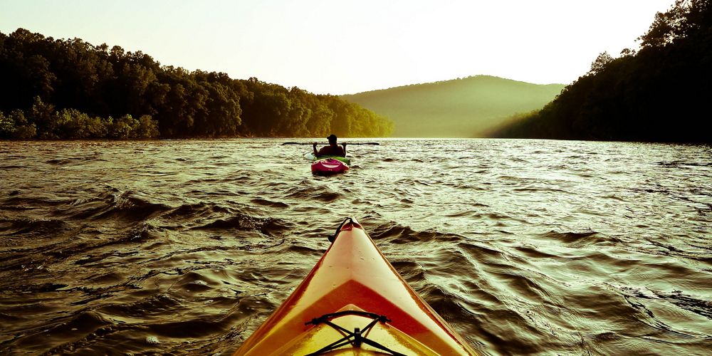 View from a kayak of another kayak on a large body of water with dense forests on all sides.
