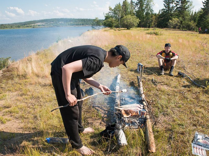 first nation teenage boy roasts marshmallow over fire next to a lake in thaidene nene