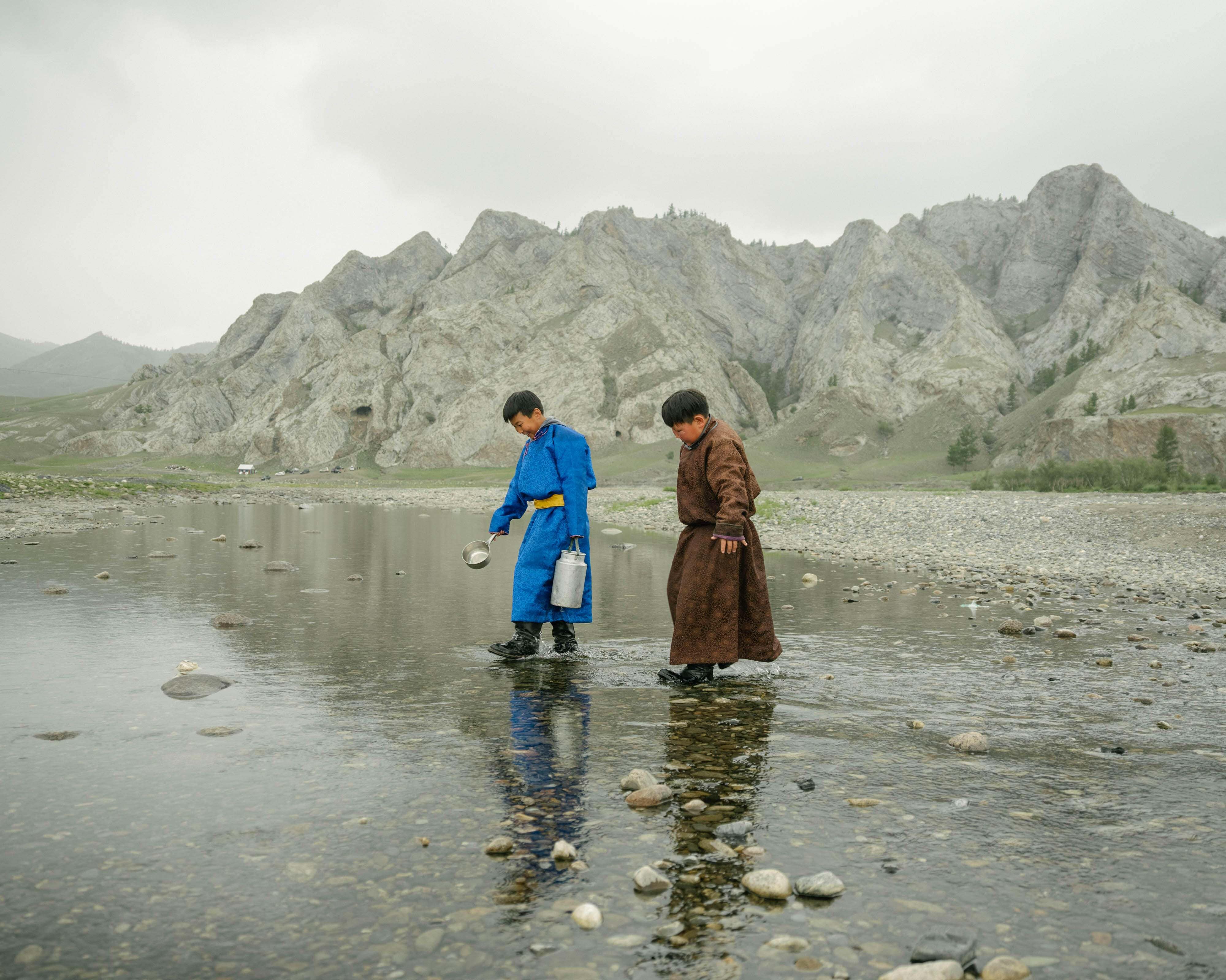 Two children walking in a body of water with mountains in the background.