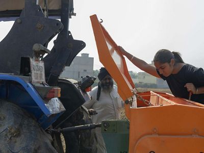 Amandeep lifts a lid from an orange seeder machine and peers inside of the machinery. Her father is nearby.